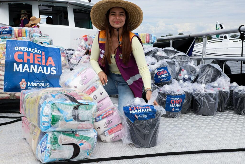 Mulher usando chapéu participa de ação social em embarcação ao lado de kits de assistência destinados a famílias em Manaus.
