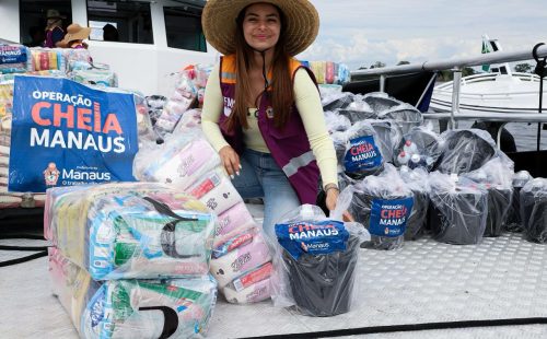 Mulher usando chapéu participa de ação social em embarcação ao lado de kits de assistência destinados a famílias em Manaus.